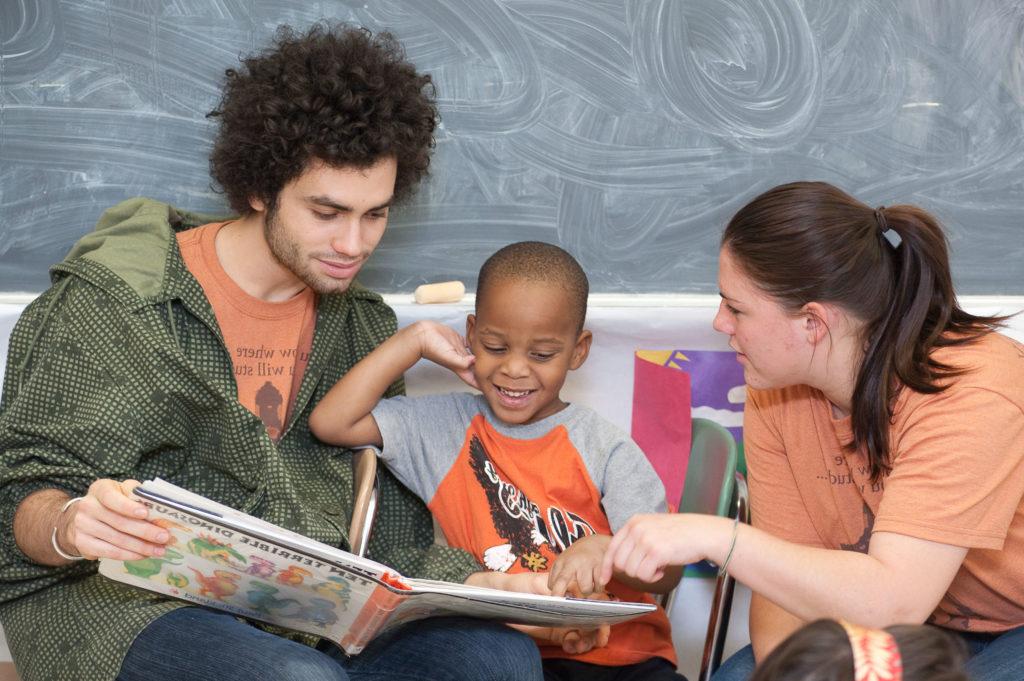 Two University of Rochester students reading a picture book to a young child in a children's classroom