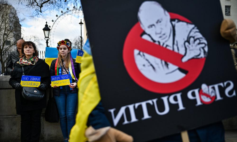 Close-up of anti-Putin rally sign with people in the background holding signs with the Ukraine flag's colors.
