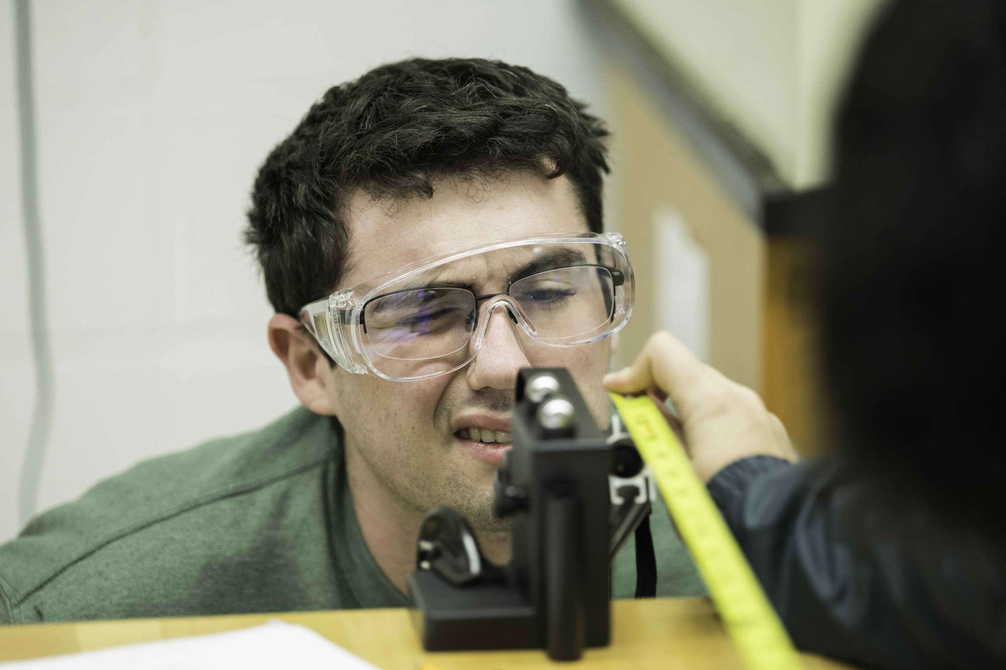 Mechanical 工程师ing student in safety goggles stares intently at equipment while a hand nearby holds measuring tape.